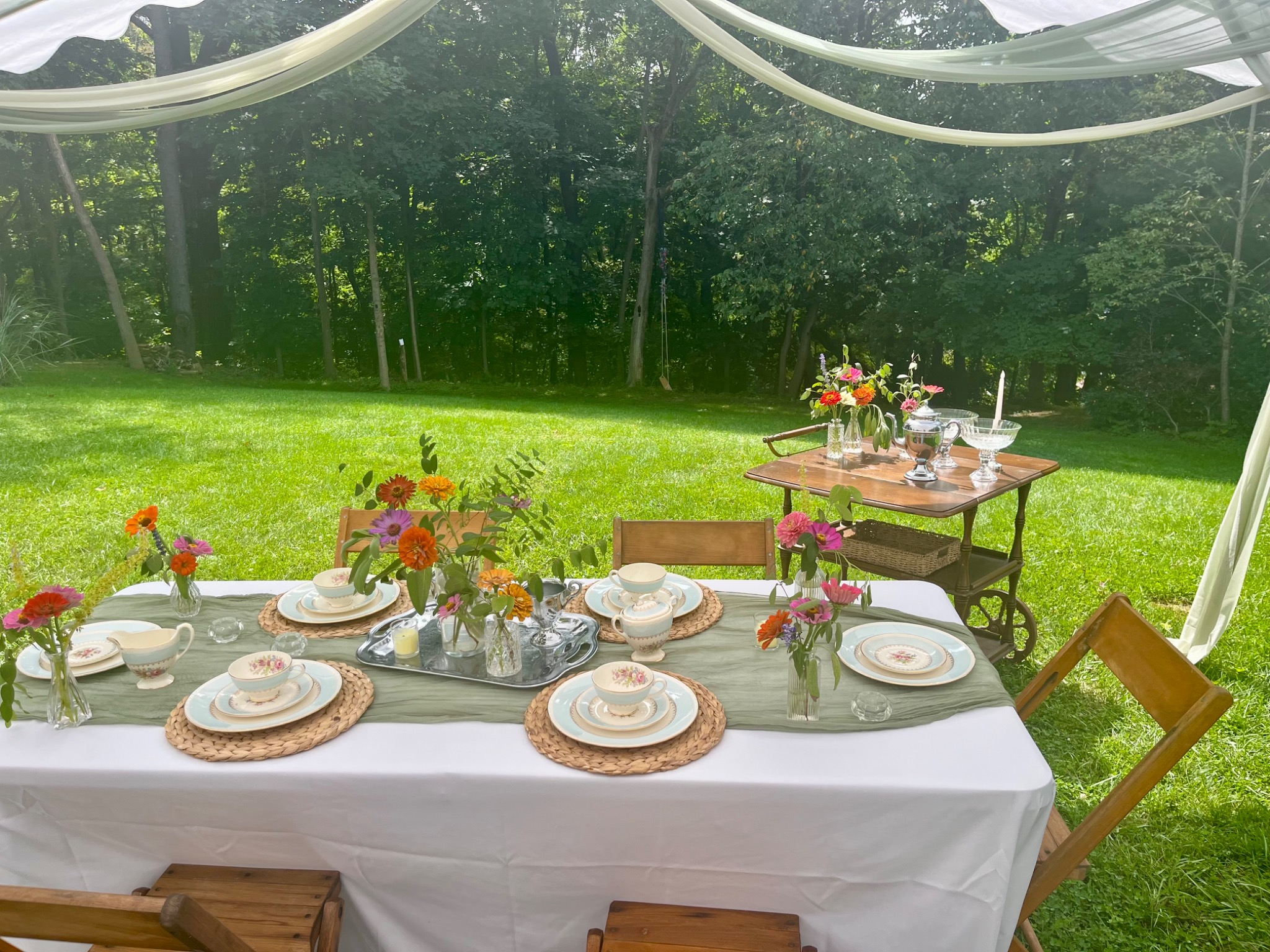 Garden tea party table with china, fresh flowers, wooden chairs and a vintage tea cart beneath a draped tent