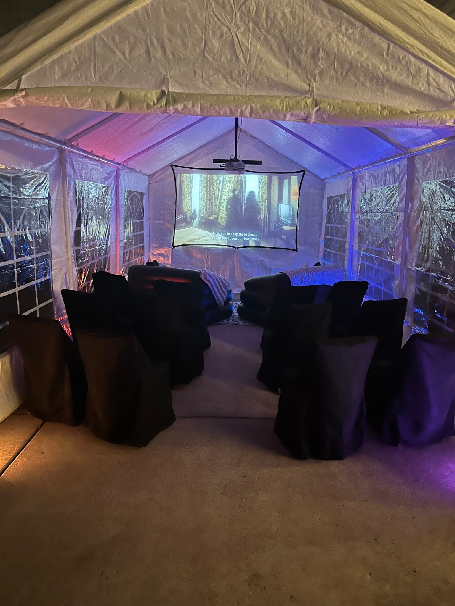 Backyard movie-night setup with a projector screen, inflatable sofas and covered chairs inside a colorfully lit tent