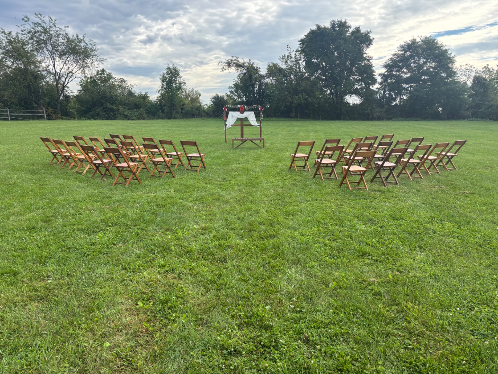 Wooden folding chairs arranged around a center aisle facing a flower-decorated wedding arbor on a lawn