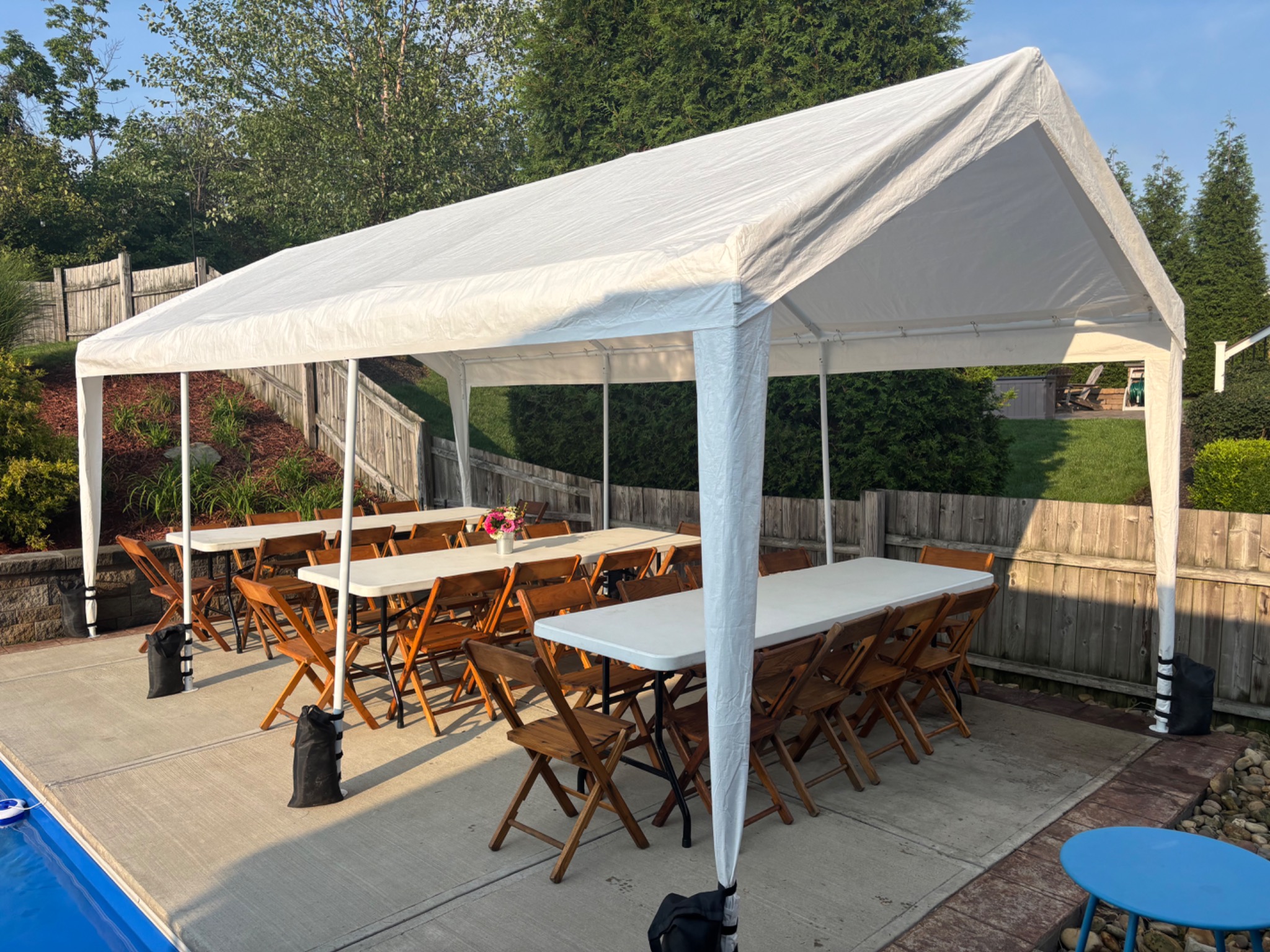 White party tent with tables, wooden folding chairs and flowers set up beside a backyard pool