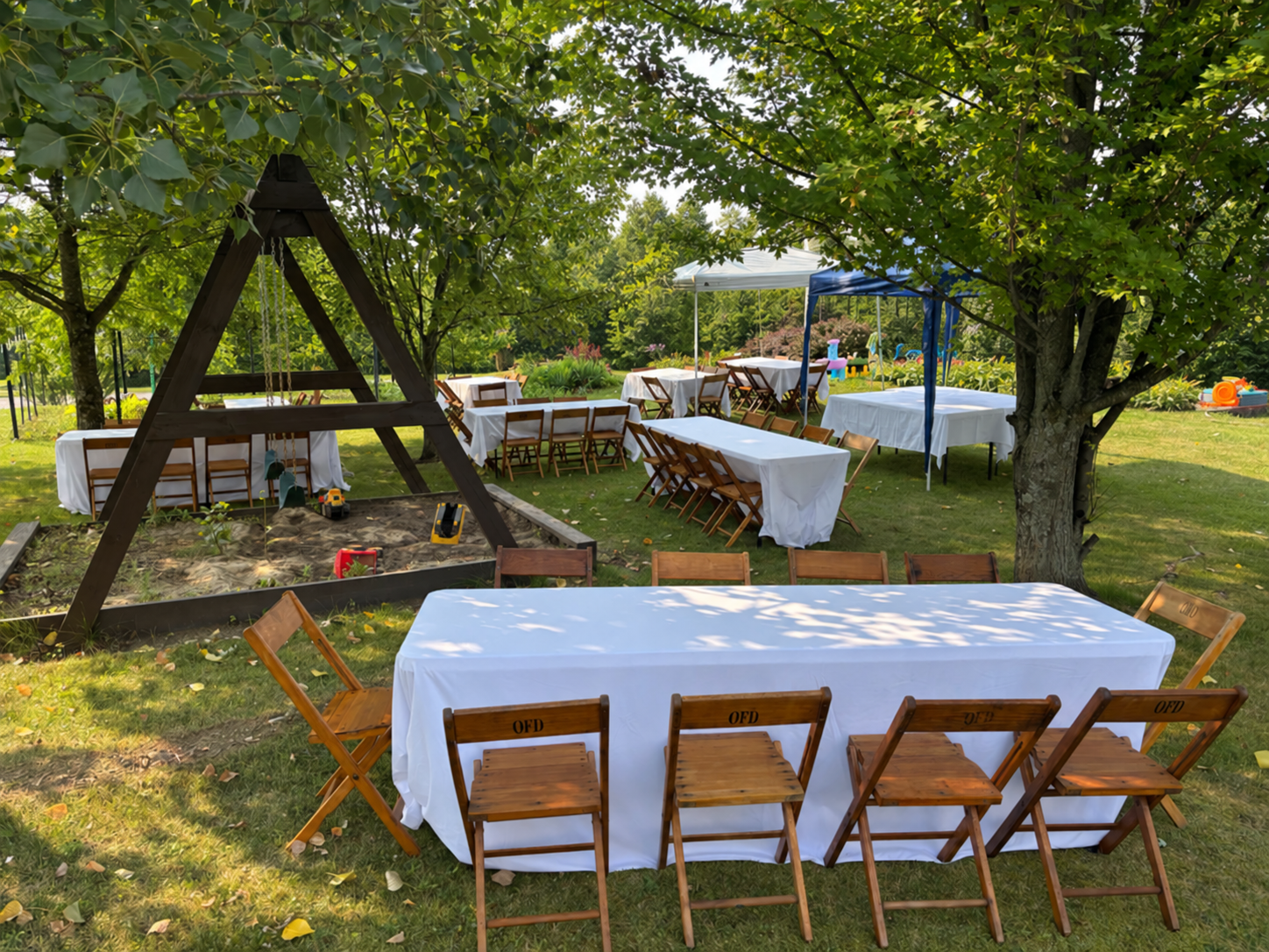 Open-air party setup with white-covered tables and wooden chairs arranged around a backyard playground